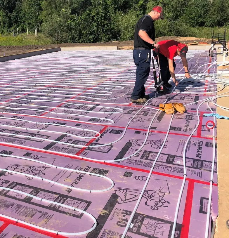 Two Comfort Heating technicians laying radiant floor heating tubing in serpentine loops across insulation board on an open slab
