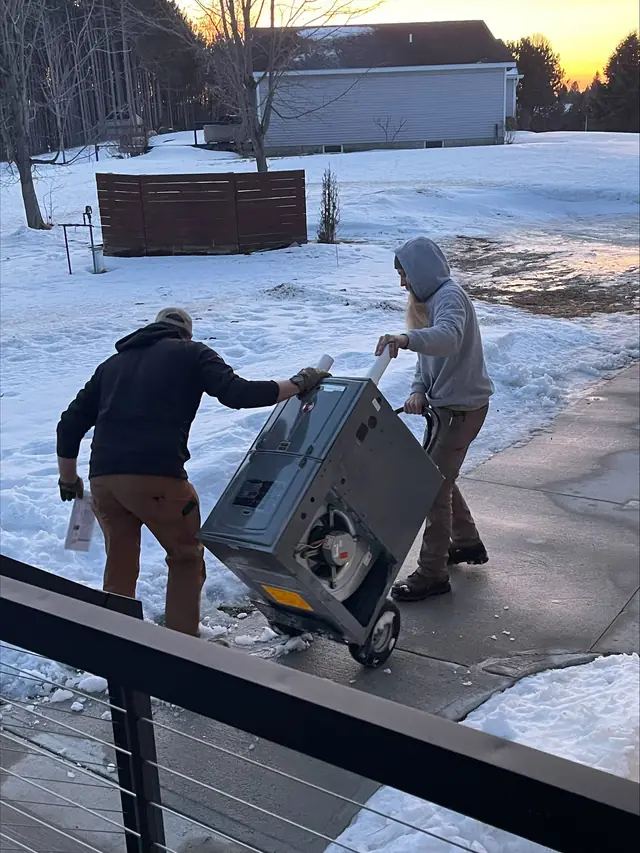 Two Comfort Heating technicians moving a furnace on a hand truck across a snowy driveway at sunset